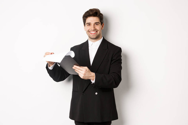 Εικόνα του αισιόδοξου επιχειρηματία με κοστούμι κοιτάζοντας τα έγγραφα, κρατώντας πρόχειρο και χαμογελώντας ευχαριστημένος, στέκεται πάνω σε λευκό φόντο - Φωτογραφία, εικόνα