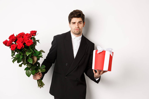 Concept of holidays, relationship and celebration. Handsome and confident man in black suit, going on a date, holding bouquet of roses and present, standing against white background - Photo, Image