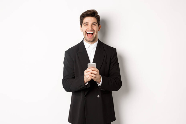 Retrato de homem feliz de boa aparência, vestindo terno preto, rindo de felicidade e usando telefone celular, em pé sobre fundo branco - Foto, Imagem