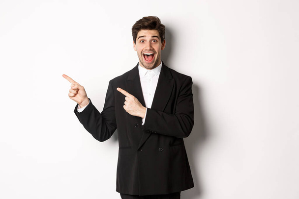 Portrait of excited and happy man in suit showing advertisement, pointing fingers at upper left corner and smiling amazed, standing over white background - Photo, image