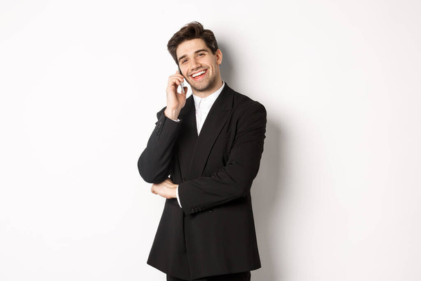 Kuva komea, onnistunut liikemies puhuu puhelimessa, hymyillen tyytyväinen, seisoo puku valkoista taustaa vasten - Valokuva, kuva