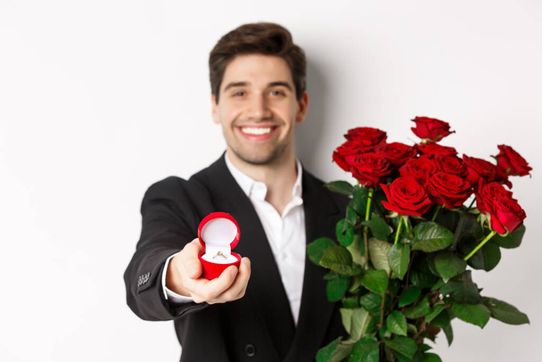 Close-up of attractive man in suit, holding bouquet of roses giving an engagement ring, proposing to girlfriend, standing against white background - Фото, зображення