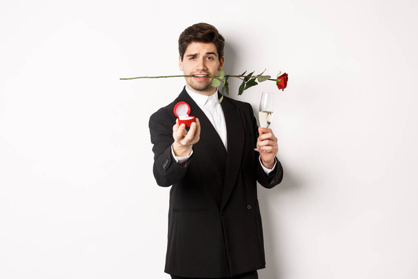 Romantic young man in suit making a proposal, holding rose in teeth and glass of champagne, showing engagement ring, asking to marry him, standing against white background - Foto, Bild