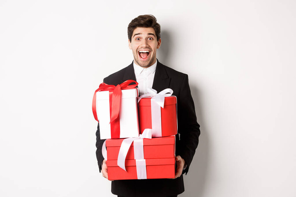 Concept of christmas holidays, celebration and lifestyle. Image of handsome amazed guy in suit, holding new year presents and smiling, standing against white background - Photo, image