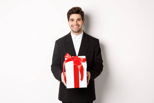 Noel tatili, kutlama ve yaşam tarzı kavramı. Siyah takım elbiseli yakışıklı bir adam, romantik bir hediyesi var, bir kutuda ve gülümsüyor, beyaz arka planda duruyor. - Fotoğraf, Görsel