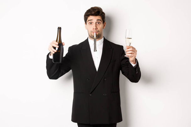 Concept of holidays, party and celebration. Portrait of handsome guy in black suit, raising bottle of champagne and glass, blowing a party whistle, having a birthday, standing over white background - Фото, изображение