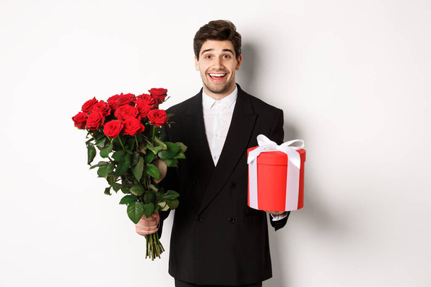 Koncept svátků, vztahu a oslav. Obrázek pohledného usmívajícího se chlapa v černém obleku, držící kytici rudých růží a dávající dárek na Nový rok, bílé pozadí - Fotografie, Obrázek
