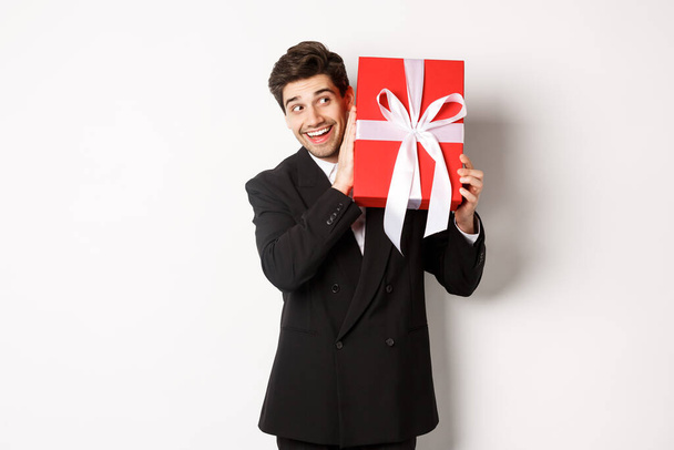Konzept von Weihnachtsurlaub, Feier und Lebensstil. Bild des aufgeregten Mannes, der das neue Jahr genießt, schüttelt Geschenkbox, um zu erraten, was drinnen vor weißem Hintergrund steht - Foto, Bild