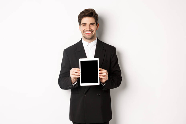 Portrait of handsome businessman in trendy suit, showing digital tablet screen and smiling, standing against white background - Фото, зображення