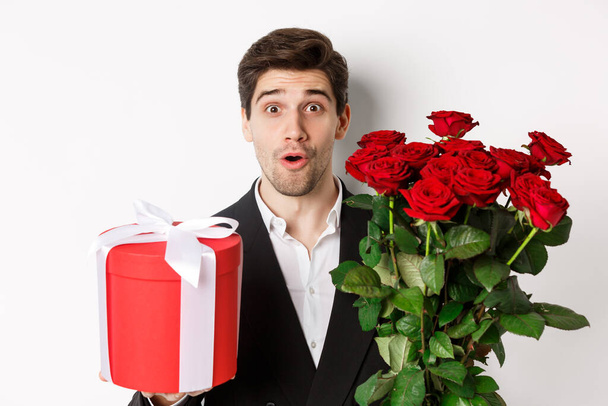 Close-up of attractive man in suit looking surprised, holding gift box and bouquet of roses, giving present for holiday, standing against white background - Photo, Image
