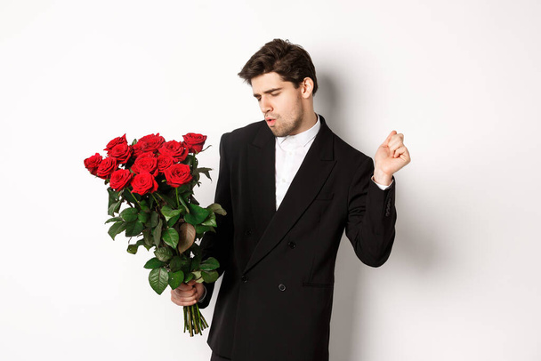Image of elegant and sassy man in black suit, looking confident and holding bouquet of red roses, going on a romantic date, standing against white background - Foto, imagen