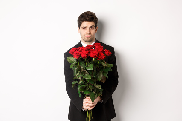 Image of handsome boyfriend in black suit, holding bouquet of red roses and smiling, being on a date, standing over white background - Zdjęcie, obraz