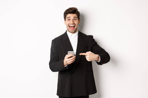 Εικόνα του χαρούμενου επιχειρηματία που αναζητούν έκπληκτος, δείχνοντας το κινητό τηλέφωνο, στέκεται στο κοστούμι πάνω από λευκό φόντο - Φωτογραφία, εικόνα