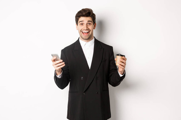 Portrait of happy good-looking man in suit, holding cup of coffee and smartphone, achieve app goal, standing over white background - Photo, Image