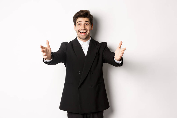 Porträt eines glücklichen attraktiven Mannes im Anzug, der eine Party ausrichtet, die Hände zur Begrüßung nach vorne streckt, etwas halten oder umarmen möchte, vor weißem Hintergrund stehend - Foto, Bild