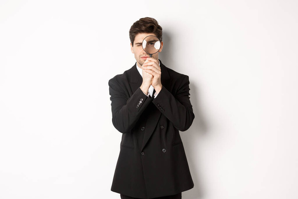 Imagem de homem de negócios sério em terno moderno preto, olhando através de lupa, à procura de funcionários, de pé contra fundo branco - Foto, Imagem