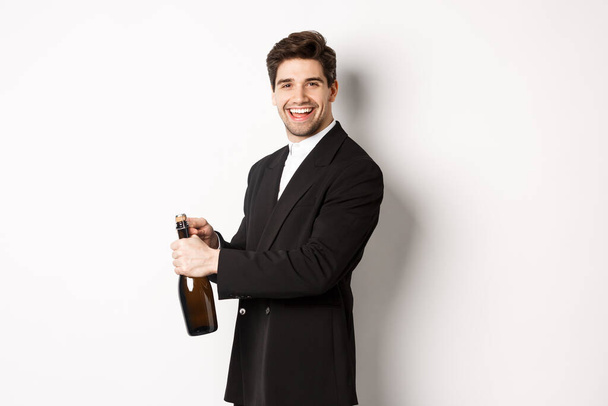 Schöner lächelnder Mann im schicken Anzug öffnet eine Flasche Champagner, feiert Feiertage, steht vor weißem Hintergrund - Foto, Bild
