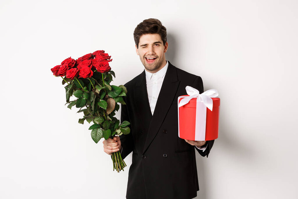 Concept of holidays, relationship and celebration. Handsome and confident man in black suit, going on a date, holding bouquet of roses and present, standing against white background - Photo, Image