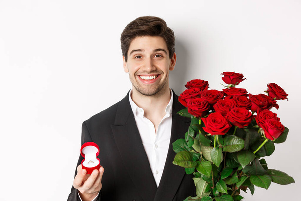 Close-up of attractive man in suit, holding bouquet of roses and engagement ring, making proposal, standing against white background - Foto, immagini
