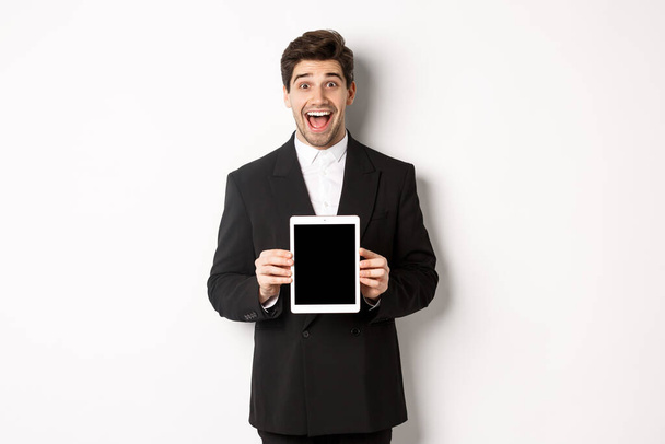 Modayı takip eden takım elbiseli çekici erkek girişimci görüntüsü, dijital tablet ekranı gösteriyor ve şaşkınlık içinde gülümsüyor, beyaz arka planda duruyor. - Fotoğraf, Görsel