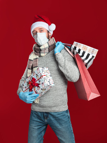 Een jonge man met een medisch masker en handschoenen, met een kerstmuts en sjaal draagt een geschenkdoos en boodschappentassen op een rode achtergrond - Foto, afbeelding