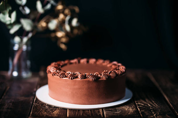 Schokoladenkuchen auf einem weißen Teller. Auf verschwommenem schwarzem Hintergrund eine Vase mit Blumen. Holztisch mit schönem Muster. Traditionelle Süßigkeiten zum Geburtstag und zu anderen Feiertagen. - Foto, Bild