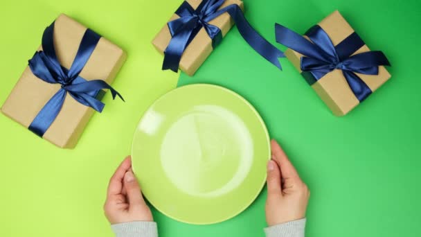 empty round ceramic green plate and female hands hold a fork with a knife on a green background with wrapped gift boxes, top view - Footage, Video