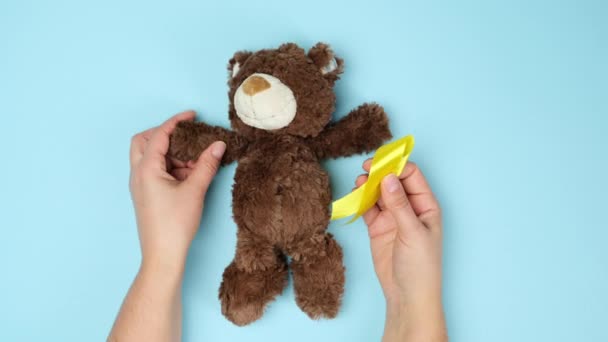 vrouwelijke handen houden een kleine teddybeer met een geel lint gevouwen in een lus op een blauwe achtergrond. het concept van de bestrijding van kinderkanker. zelfmoordprobleem. bovenaanzicht - Video