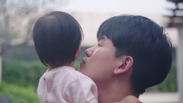 Head shot of young Asian father holding and kissing his adorable cute baby girl, small kid child care parenting, standing outside shot, Family member bonding, learning abilities in childhood, family tenderness, father and daughter happy scene - Footage, Video