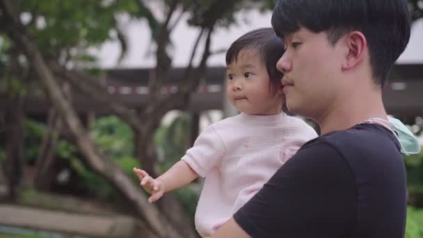 Asyalı sevimli küçük kız babasının kollarında, parkta bebeği tutan genç ebeveyn, çocuk bakımı, Asyalı aile üyesi, mutluluk bağı ve ilişki, çocukluk öğrenme yeteneği, çocukta masumiyet merakı - Video, Çekim