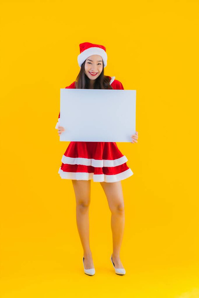 Porträt schöne junge asiatische Frau Weihnachten Kleidung und Hut Lächeln mit leeren weißen Brett Zeichen auf gelbem Hintergrund isoliert - Foto, Bild