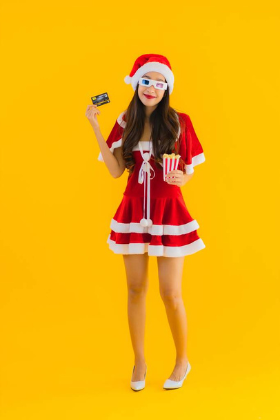 ポートレート美しい若いアジアの女性は、ポップコーンと3Dメガネでクリスマス服の帽子を身に着けています準備ができた時計の映画のために黄色隔離された背景 - 写真・画像