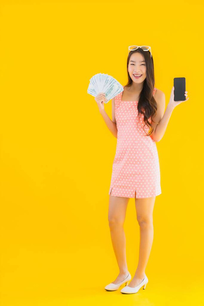 Porträt schöne junge Asiatin zeigt viel Bargeld und Geld mit Handy auf gelbem Hintergrund - Foto, Bild