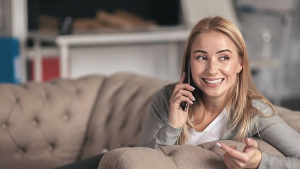 Beautiful young woman talking on the phone while sitting at home on the sofa - Foto, imagen