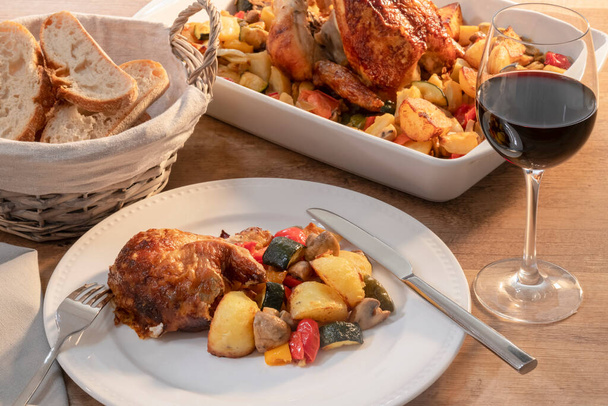 a plate of chicken and vegetables surrounded by a basket of bread, a glass of wine and a baking dish - Photo, image