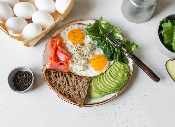 Gebakken eieren op wit bord met avocado, brood en sla op een lichte achtergrond. Bovenaanzicht. Kopieerruimte - Foto, afbeelding