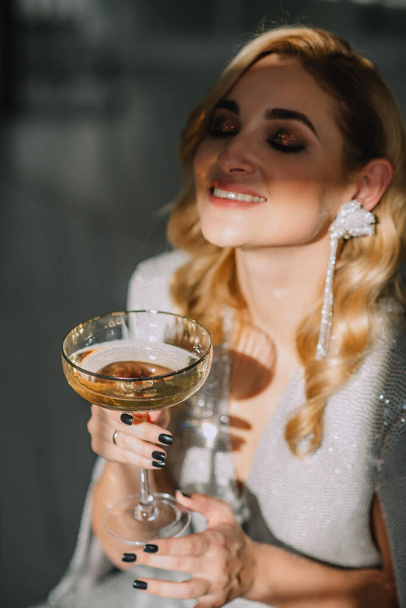 Young beautiful luxurious blonde woman with a glass in her hands celebrates the New Year against the background of blurry lights bokeh of a Christmas tree. Soft selective focus, art noise. - Foto, immagini