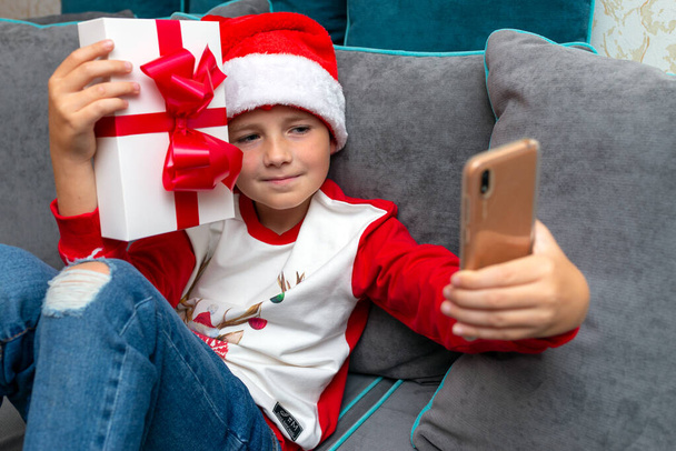 Handsome kid boy in Santa Claus hat looks at smartphone screen, shows a christmas present.Talk with family,closeup image. - Photo, Image