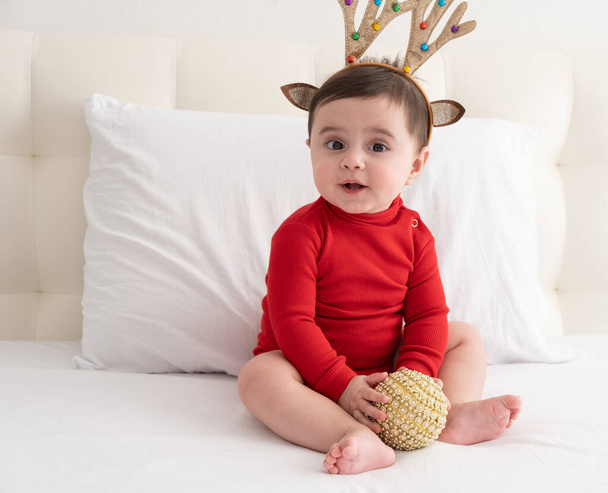 κοριτσάκι 6 μηνών σε κόκκινο κοστούμι του σώματος με κορδέλα με κέρατα ελαφιού κάθεται σε λευκό κρεβάτι. - Φωτογραφία, εικόνα