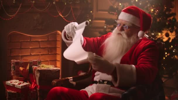 Літній чоловік, одягнений як Санта Клаус, читає список побажань у кімнаті на Різдво, тримаючи довгий білий аркуш в руках. - Кадри, відео