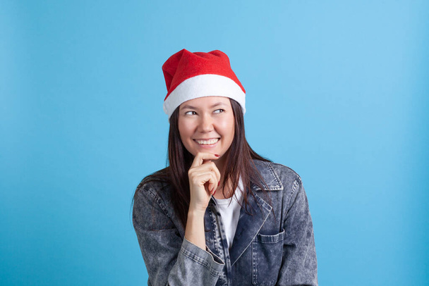 Noel Baba şapkası takmış gülümseyen Asyalı genç bir kadın. Çenesini tutuyor ve mavi bir arka planda tek başına gözlerini kaçırıyor. - Fotoğraf, Görsel