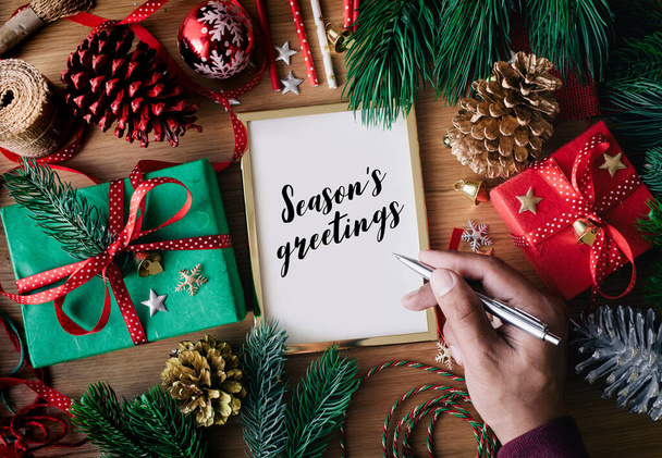 Human hand writing Season's greeting text on cards with gift box present and ornament element on wood table background.winter season activity ideas - Photo, Image