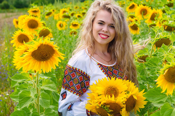 Belle fille blonde ukrainienne aux cheveux longs portant une chemise ethnique nationale parmi les tournesols. Jeune femelle souriante tenant un bouqet de tournesols et se tenant au milieu d'un champ en fleurs. - Photo, image