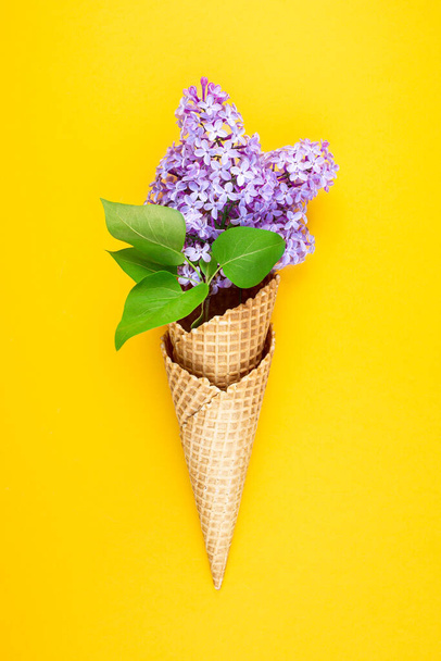 Ice cream cone with Lilac flowers on yellow background. Waffle cup with spring flowers. Greeting card. Minimalism fashion style. Copy space, flat lay - Photo, Image