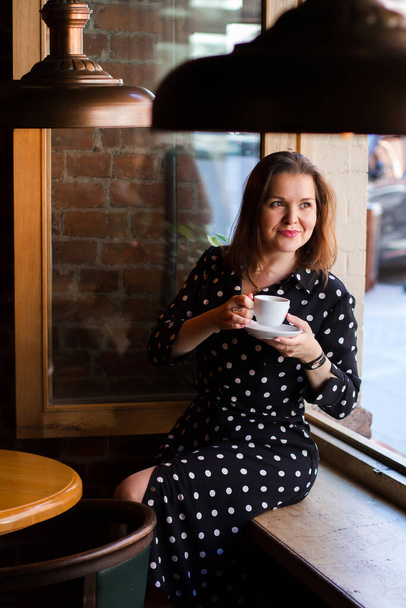 Portrait of beautiful woman drinking a cup of coffee while sitting near the window in modern loft cafe bar. Portrait of dreaming lady looking away. - Fotó, kép