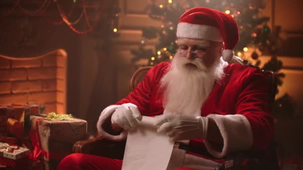 Kerstman zit in zijn gezellige kamer in residentie met kerstvakantie, rollende lijst met wensen, leuke oude man  - Video