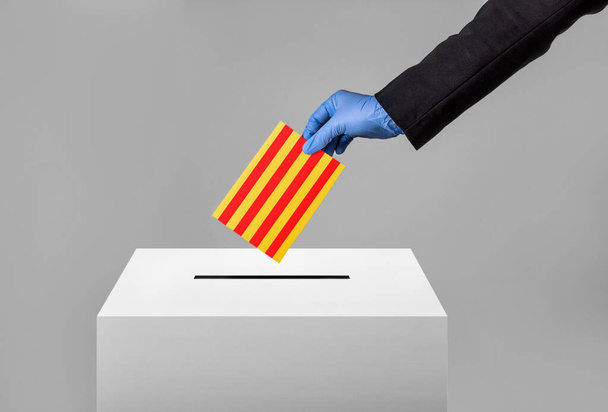 Een hand met een beschermhandschoen die een enveloppe met de Catalaanse vlag in de stembus stopt voor de Catalaanse verkiezingen van 2021 in de tijd van covid-19, met een grijze achtergrond. - Foto, afbeelding