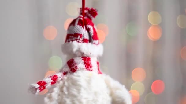 Hermoso juguete árbol de Navidad cuelga en el fondo de luces de Navidad multicolores y se mueve ligeramente. Primer plano. El juguete del árbol de Navidad en forma del muñeco de nieve de Año Nuevo. - Imágenes, Vídeo