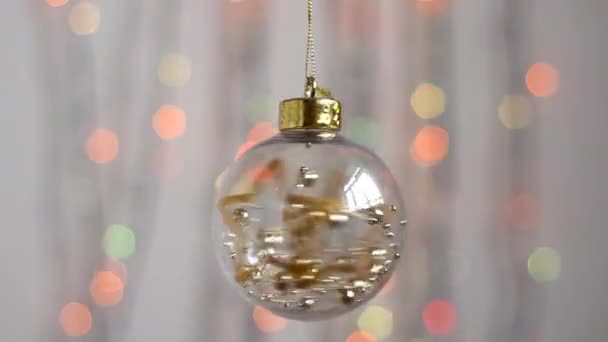 Schöne Weihnachtsbaumspielzeug hängt vor dem Hintergrund der bunten Weihnachtsbeleuchtung und bewegt sich leicht. Nahaufnahme. Transparentes Weihnachtsbaumspielzeug in Form einer Kugel mit goldenen Sternen und Kugeln im Inneren. - Filmmaterial, Video