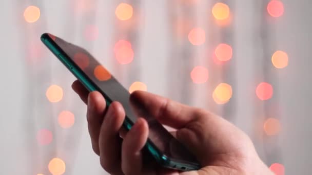 Man is typing SMS on his mobile phone against the background of New Year's lights. A young man makes an online order by phone during quarantine. Online shopping, New Year's gifts, Christmas flare. - Footage, Video